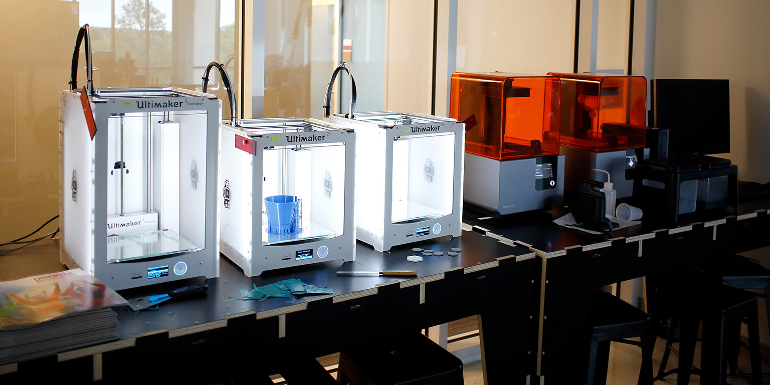 Five 3D printers lined up in a row on a table next to a lab desktop computer