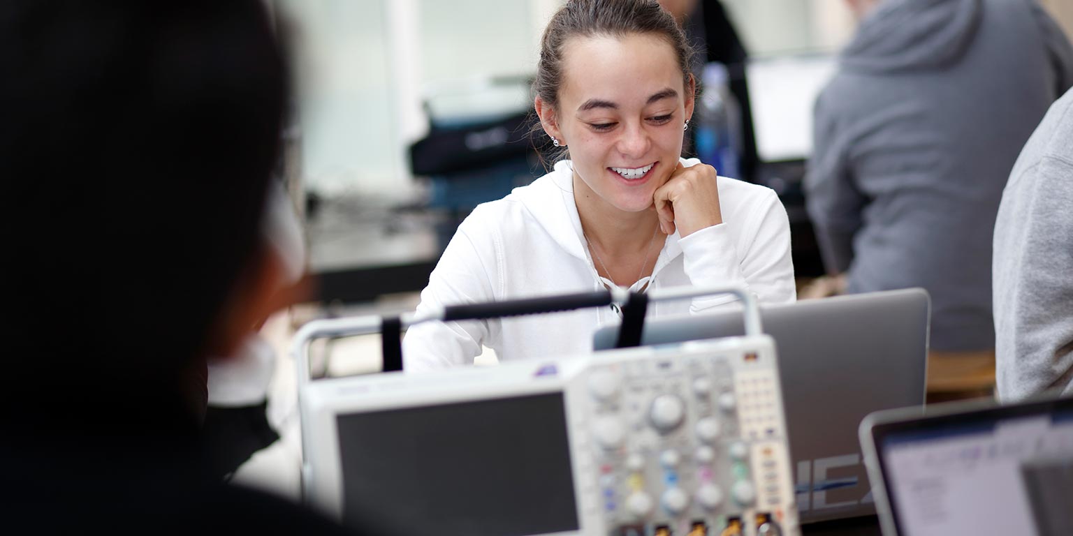 A student smiles as she works on her computer in a lab with other students. 