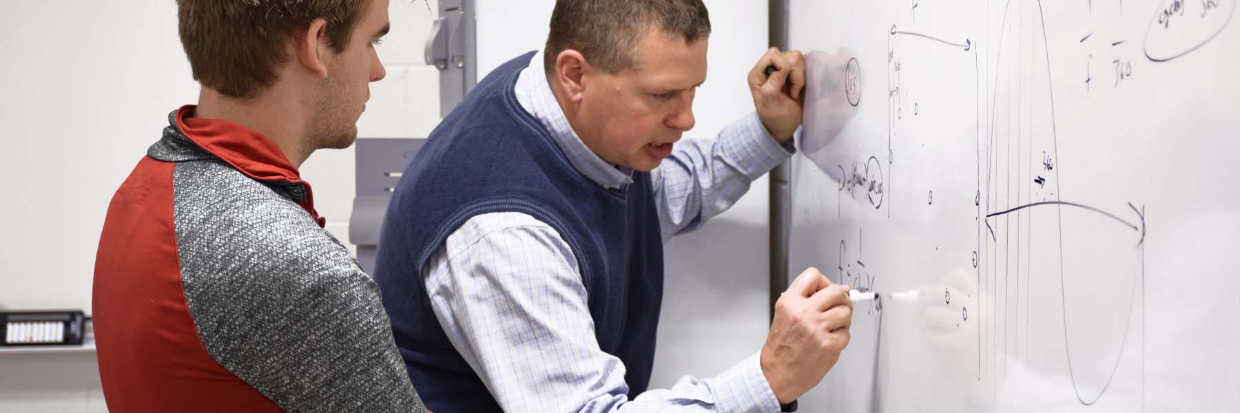 A professor writes an equation on a whiteboard as a student watches. 