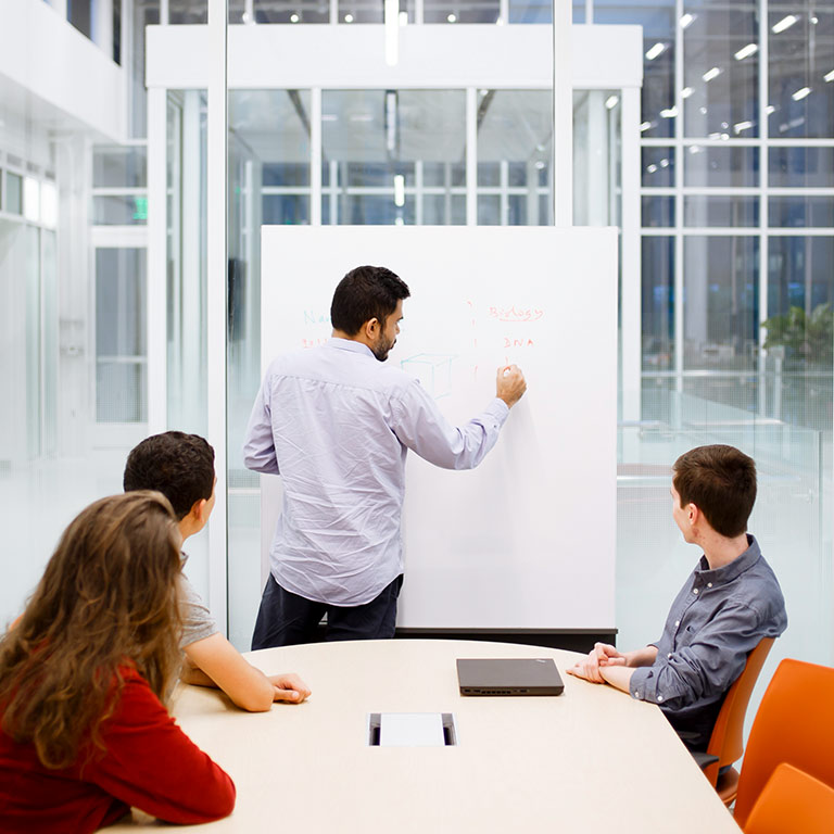 Students sitting at a table watch a professor write on a whiteboard in a Luddy Hall classroom.